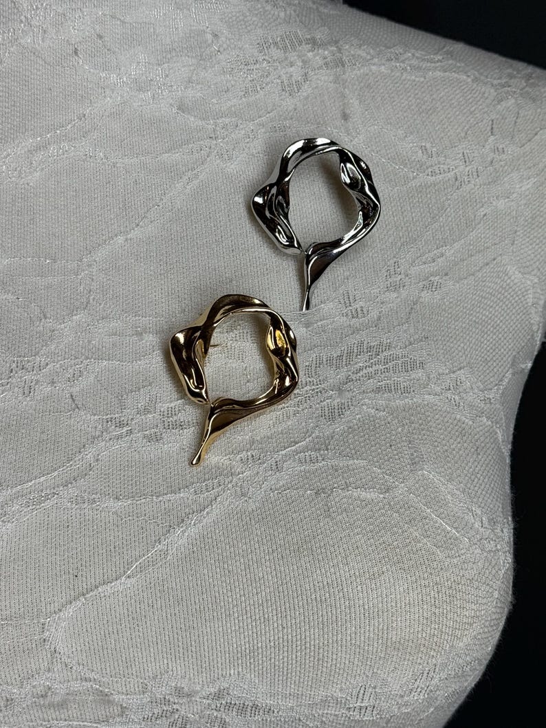May include: Two abstract metal rings, one gold and one silver, with a wavy, organic design. The rings are displayed on a white fabric background.