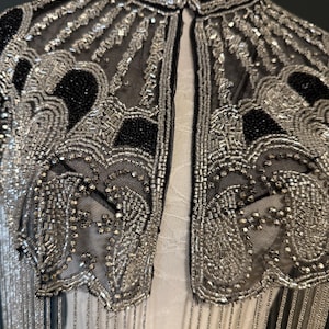 May include: Close-up of a black and silver beaded jacket. The jacket features intricate silver beadwork on a sheer black fabric, with a high collar and fringe detail. The design includes geometric and floral patterns, creating a vintage aesthetic.