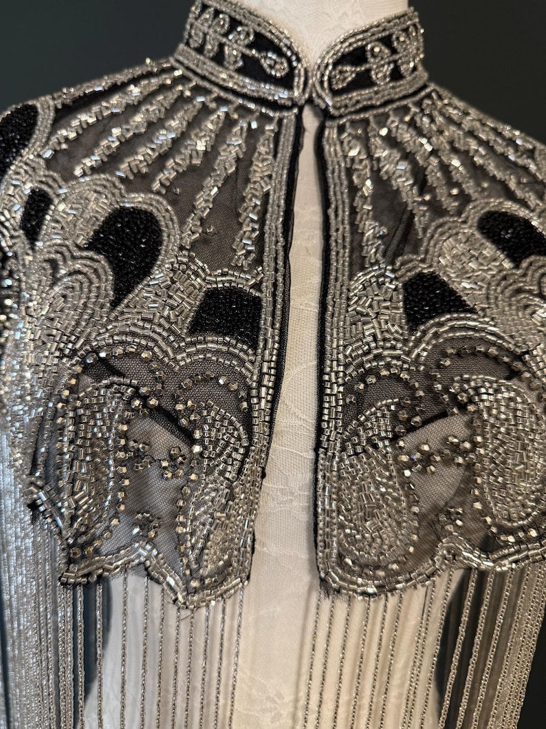 May include: Close-up of a black and silver beaded jacket. The jacket features intricate silver beadwork on a sheer black fabric, with a high collar and fringe detail. The design includes geometric and floral patterns, creating a vintage aesthetic.