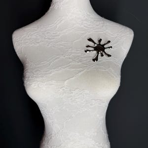 Puede incluir: Un broche plateado en forma de estrella estilizada. El broche est&aacute; sujeto a un maniqu&iacute; de tela blanca.
