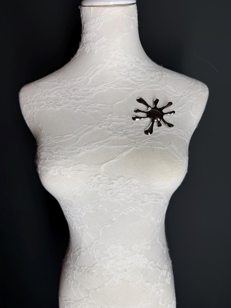 Puede incluir: Un broche plateado en forma de estrella estilizada. El broche est&aacute; sujeto a un maniqu&iacute; de tela blanca.