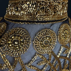 May include: Close-up of a black garment with detailed gold beadwork. The design features a high, embellished collar and patterns of gold beads on a sheer fabric, creating a luxurious and ornate appearance. The beadwork includes geometric and floral motifs.