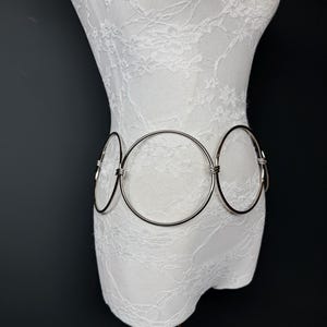 Może przedstawiać: Srebrny pasek metalowy z trzema połączonymi kółkami. Pasek znajduje się na białym manekinie pokrytym koronką.