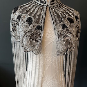 May include: A silver beaded cape with black accents and fringe. The cape features intricate beadwork and a high collar. The design includes a sheer mesh fabric and long, dangling fringe.