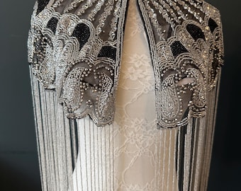 May include: A silver beaded cape with black accents and fringe. The cape features intricate beadwork and a high collar. The design includes a sheer mesh fabric and long, dangling fringe.