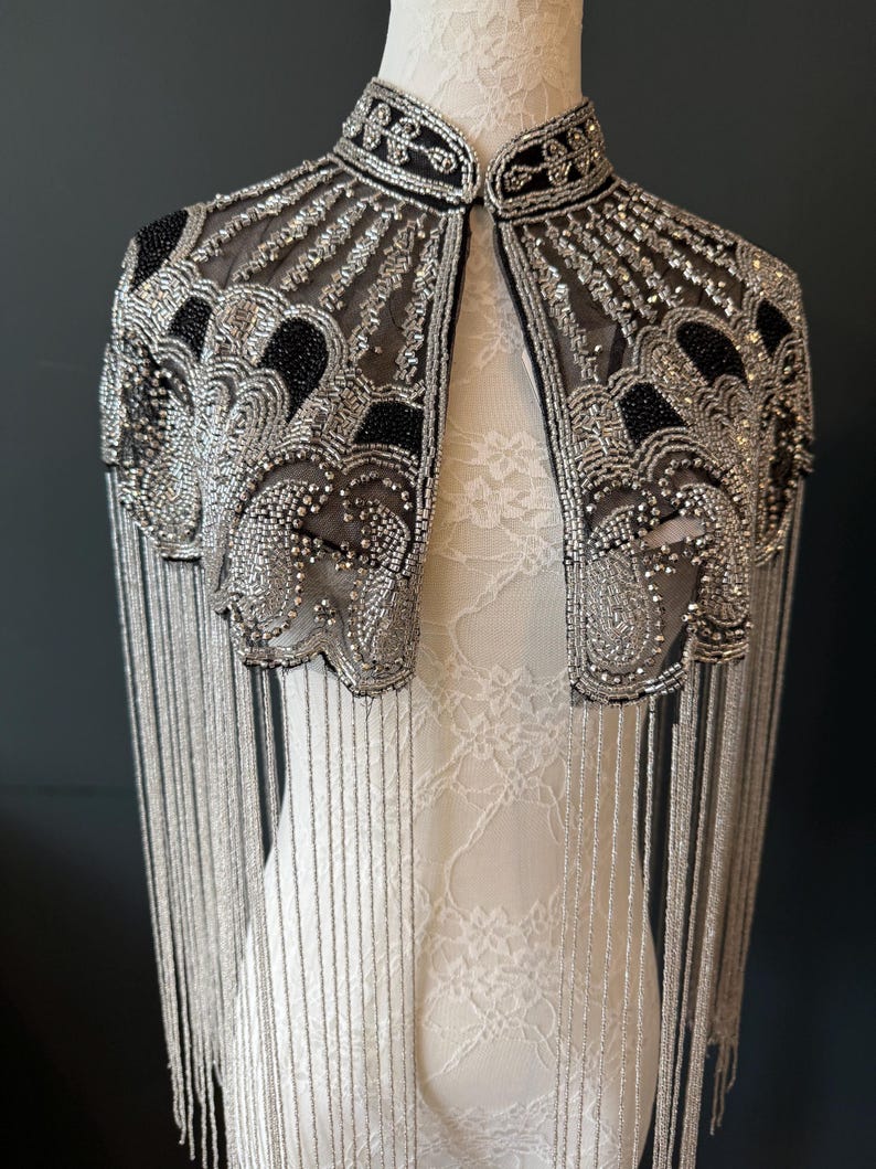 May include: A silver beaded cape with black accents and fringe. The cape features intricate beadwork and a high collar. The design includes a sheer mesh fabric and long, dangling fringe.