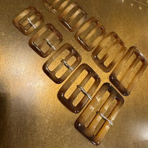 Puede incluir: Un juego de ocho hebillas de cinturón de plástico marrón con un patrón de mármol. Las hebillas tienen un cierre de metal plateado.