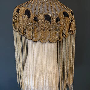 May include: A gold and black beaded cape with fringe. The cape features intricate beadwork in a floral pattern around the neckline and shoulders. Long gold fringe hangs from the bottom edge, creating a dramatic effect. The cape is displayed on a mannequin.