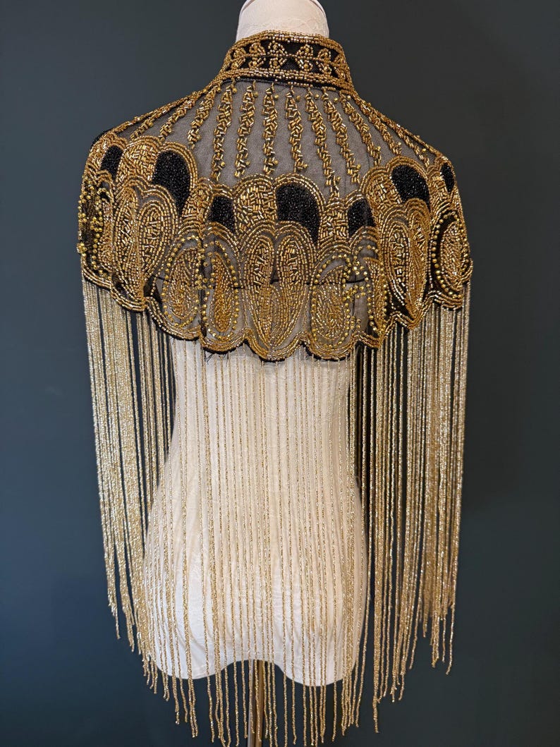 May include: A gold and black beaded cape with fringe. The cape features intricate beadwork in a floral pattern around the neckline and shoulders. Long gold fringe hangs from the bottom edge, creating a dramatic effect. The cape is displayed on a mannequin.