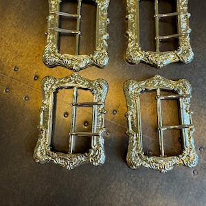 May include: Four ornate, gold-colored metal buckles with decorative scrollwork and a rectangular shape. Each buckle has two horizontal bars for securing straps. The buckles are arranged on a dark, textured surface.