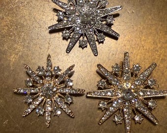 May include: Three starburst brooches with crystal-like embellishments. One brooch is silver-toned, while the other two are gold-toned. Each brooch has a large central stone and smaller stones on the points.