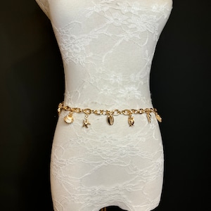 Puede incluir: Cinturón de cadena dorada con varios abalorios dorados en forma de conchas y una estrella de mar. El cinturón está en un maniquí cubierto de encaje blanco.
