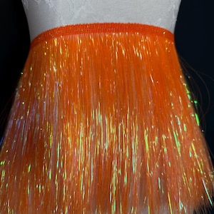 Könnte beinhalten: Leuchtend orangefarbene Fransenborte mit schimmernden Highlights. Die Borte ist an einem schmalen orangefarbenen Band befestigt. Die Fransen bestehen aus langen, dünnen Strähnen, die einen strukturierten, schimmernden Effekt erzeugen. Ideal für Kostüme oder Bastelprojekte.
