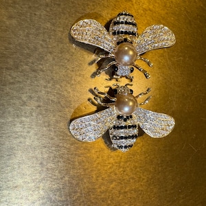 Puede incluir: Dos broches de abeja de oro con rayas blancas y negras y pedrería transparente. Cada abeja tiene una perla en el centro.
