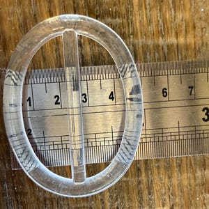 Op de afbeelding: Een doorzichtige, ovale plastic gesp met een centrale staaf, ongeveer 7,6 cm lang, ligt op een metalen liniaal. De gesp heeft subtiele patroondetails. De liniaal toont metingen in cm.