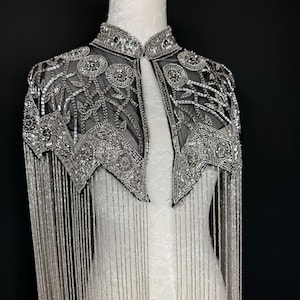 May include: A silver beaded cape with a high collar and intricate floral patterns. The cape features long, dangling fringe and is displayed on a white mannequin against a black backdrop. The cape is a formal wear item.