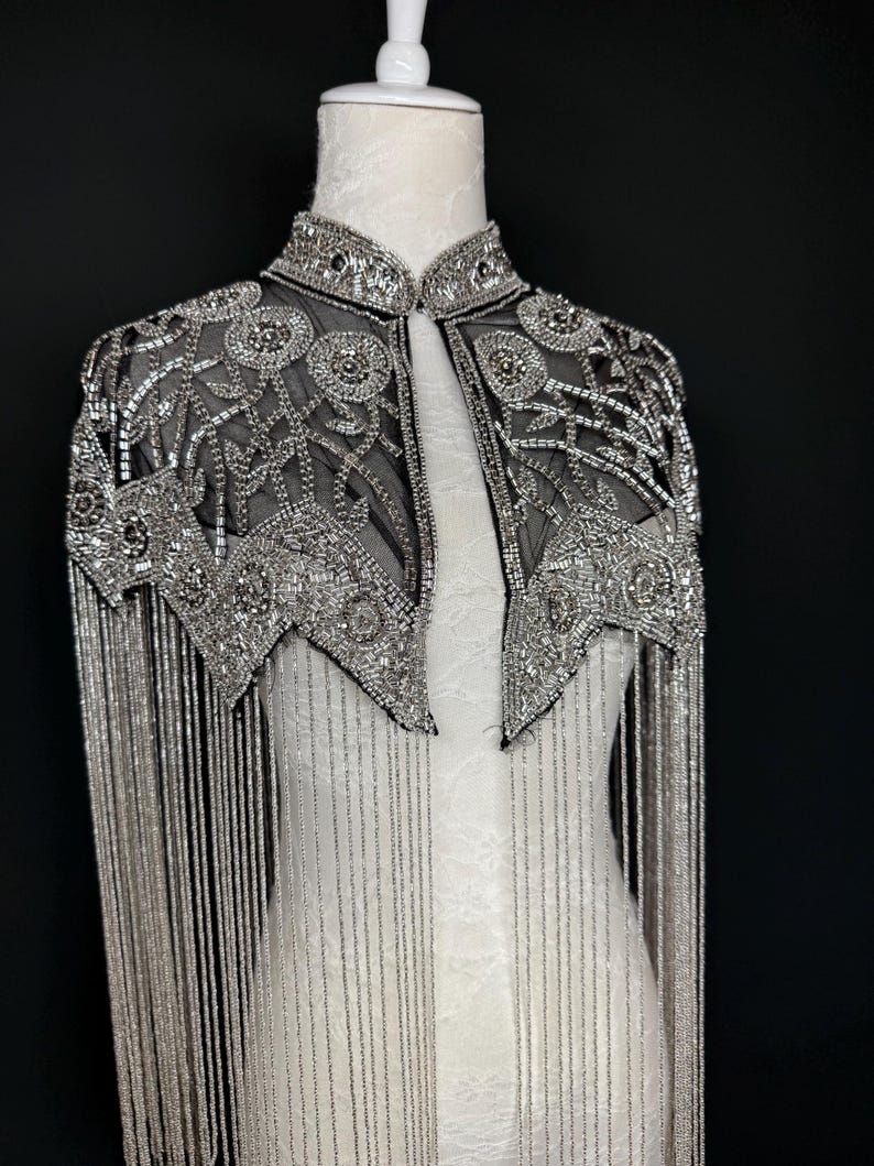 May include: A silver beaded cape with a high collar and intricate floral patterns. The cape features long, dangling fringe and is displayed on a white mannequin against a black backdrop. The cape is a formal wear item.