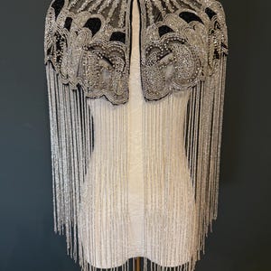 May include: A beaded cape with a high collar, featuring intricate silver and black embellishments. Long silver fringe hangs from the shoulders. The cape is displayed on a white dress form against a dark background.