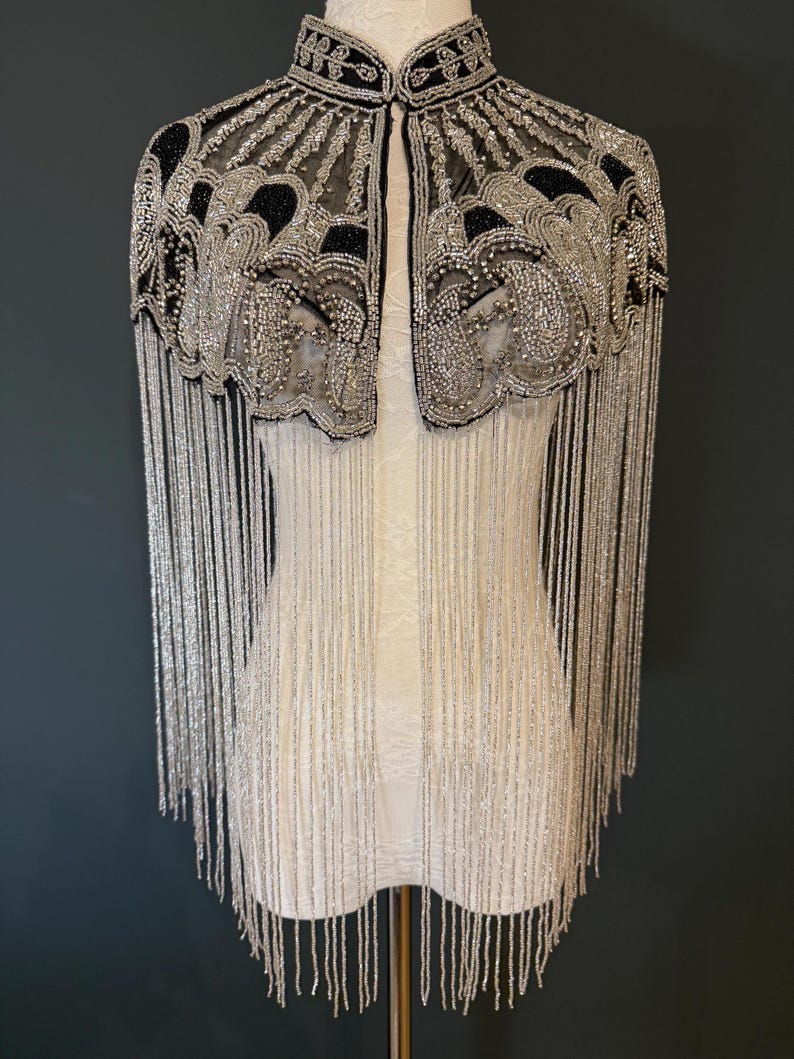 May include: A beaded cape with a high collar, featuring intricate silver and black embellishments. Long silver fringe hangs from the shoulders. The cape is displayed on a white dress form against a dark background.