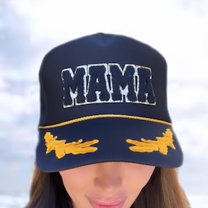 May include: A black mesh trucker hat with a gold trim and the word "MAMA" embroidered in white and black letters. The hat has gold embroidered wings on the sides.