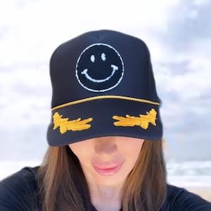 May include: A black trucker hat with a yellow brim and a white smiley face embroidered on the front. The hat also has two yellow wings embroidered on the brim.