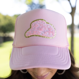 May include: A pink trucker hat with a white and gold embroidered cowboy hat design.