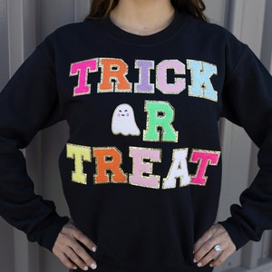 Op de afbeelding: Zwart sweatshirt met de tekst "TRICK OR TREAT" in kleurrijke letters met gouden rand en een witte geest met een roze gezicht.