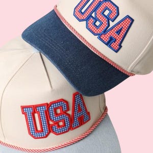 May include: Two baseball caps with the word "USA" embroidered in red and blue checkered letters. The caps have a cream-colored crown, a denim blue brim, and a red and white rope detail. The background is a soft pink.