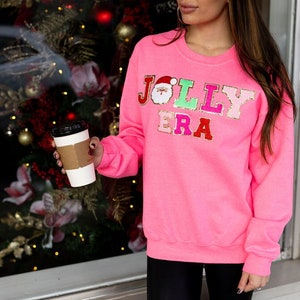 May include: A pink sweatshirt with the words "JOLLY ERA" in colorful, glittery letters. The letters are decorated with festive holiday icons, including a Santa Claus face.