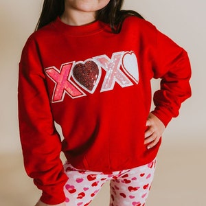 May include: A red sweatshirt with a sparkly pink and white "XOXO" graphic. The sweatshirt is worn by a child wearing pink and white heart patterned leggings and white heart-shaped sunglasses.