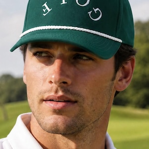 May include: A dark green golf cap with the word "GOLF" embroidered in white. The cap has a white rope detail along the brim. The background shows a golf course.