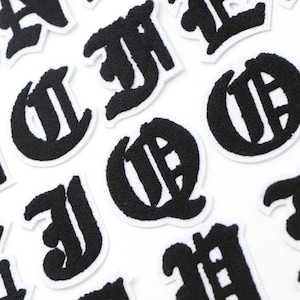 Op de afbeelding: Een set zwarte vilt strijkpatches in de vorm van oude Engelse letters. De letters zijn op een witte achtergrond.