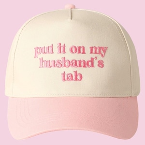 Pode incluir: Um boné de beisebol bege e rosa com o texto "put it on my husband's tab" bordado em rosa.