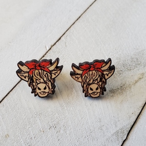 May include: A pair of wooden earrings featuring a Highland cow design. The cows are brown with a red bandana and have a white face.