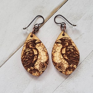 Puede incluir: Un par de pendientes de madera con un diseño grabado con láser de un cuervo y un patrón de relojería de inspiración steampunk. Los pendientes tienen forma de lágrima y tienen un gancho de color cobre.