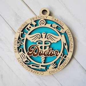 May include: A wooden ornament with a blue background and a medical symbol in the center. The ornament is decorated with medical symbols and the word "Doctor" in a brown font. The words "Loving, Friendly, Caring, Dependable, Compassionate, Comforting, Patient" are written around the edge of the ornament.