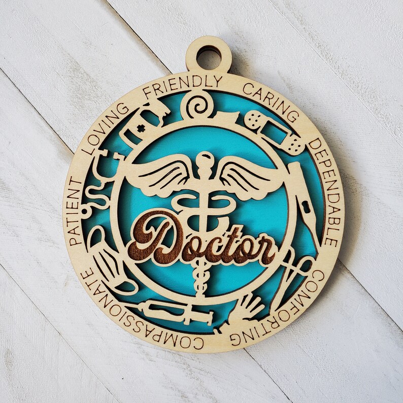 May include: A wooden ornament with a blue background and a medical symbol in the center. The ornament is decorated with medical symbols and the word "Doctor" in a brown font. The words "Loving, Friendly, Caring, Dependable, Compassionate, Comforting, Patient" are written around the edge of the ornament.