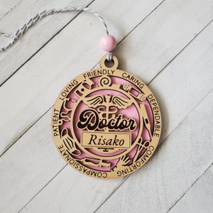 May include: A pink and brown wooden ornament with the word "Doctor" and the name "Risako" inscribed. The ornament is decorated with medical symbols and the words "patient, loving, friendly, caring, dependable, comforting, compassionate".