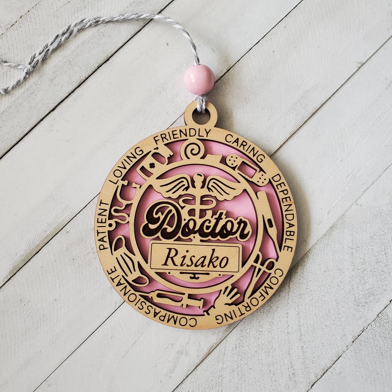 May include: A pink and brown wooden ornament with the word "Doctor" and the name "Risako" inscribed. The ornament is decorated with medical symbols and the words "patient, loving, friendly, caring, dependable, comforting, compassionate".