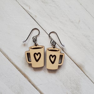 May include: A pair of wooden earrings shaped like coffee mugs with a heart design. The earrings are brown and have silver hooks.
