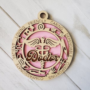 May include: A pink and brown wooden ornament with the word "Doctor" in the center. The ornament is decorated with medical symbols and the words "Patient, Loving, Friendly, Caring, Dependable, Comforting, Compassionate".
