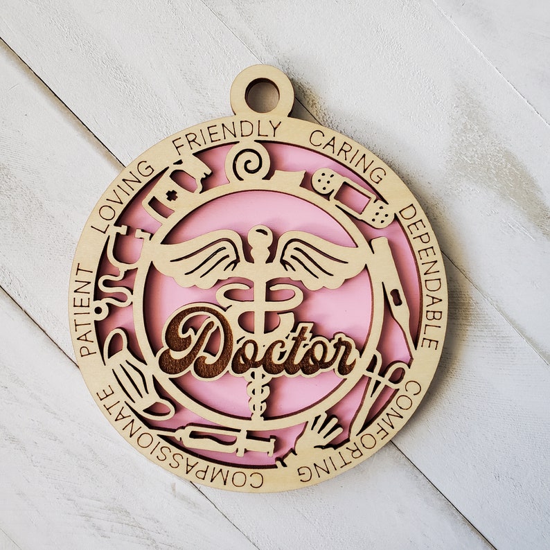 May include: A pink and brown wooden ornament with the word "Doctor" in the center. The ornament is decorated with medical symbols and the words "Patient, Loving, Friendly, Caring, Dependable, Comforting, Compassionate".