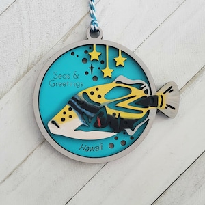 May include: A turquoise blue wooden ornament with a yellow, black, and white fish design. The ornament has the text "Seas & Greetings" and "Hawaii" on it. There are five yellow stars on the ornament.