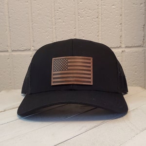Puede incluir: Una gorra de camionero de malla negra con un parche de cuero marrón de la bandera estadounidense.