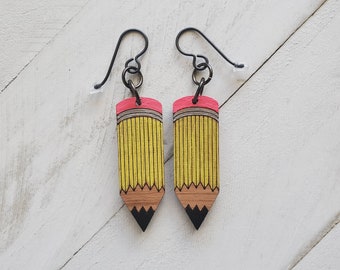 Puede incluir: Un par de pendientes de madera con forma de lápiz con un diseño de lápiz amarillo y marrón y una goma de borrar rosa. Los pendientes tienen ganchos de metal negro.