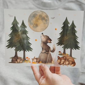 May include: A whimsical illustration featuring a wolf howling at a full moon, with two sleeping wolf pups and a pair of evergreen trees. The artwork has a soft, watercolor style with autumnal leaves and bats. The image is held over a grey sweatshirt.