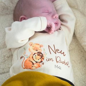 Op de afbeelding: Een crèmekleurige baby romper met een leeuw en welp illustratie en de tekst "Neu im Rudel Mats".