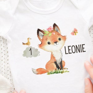 Puede incluir: Un body blanco para bebé con una ilustración de acuarela de un zorro con una corona de flores. El zorro está sentado en un parche de hierba verde. Un pequeño pájaro está volando sobre una nube gris con el número 1 en ella. Una mariposa está volando en la esquina superior derecha. El body tiene el nombre Leonie impreso en él.