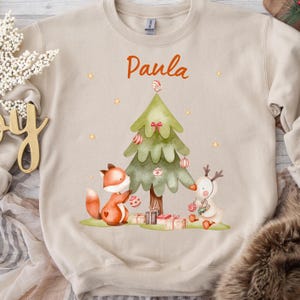May include: Beige sweatshirt with a Christmas-themed graphic. The design features a green Christmas tree, a fox, and a reindeer, all decorated with ornaments. The name "Paula" is written above the tree in orange script.