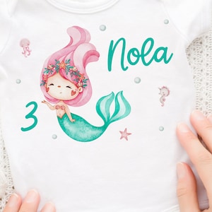 Könnte beinhalten: Weißer Strampler mit einer Meerjungfrauen-Illustration. Die Meerjungfrau hat rosa Haare, einen grünen Schwanz und trägt einen Muschelbh. Der Strampler hat auch die Zahl 3 und den Namen "Nola" in Grün.